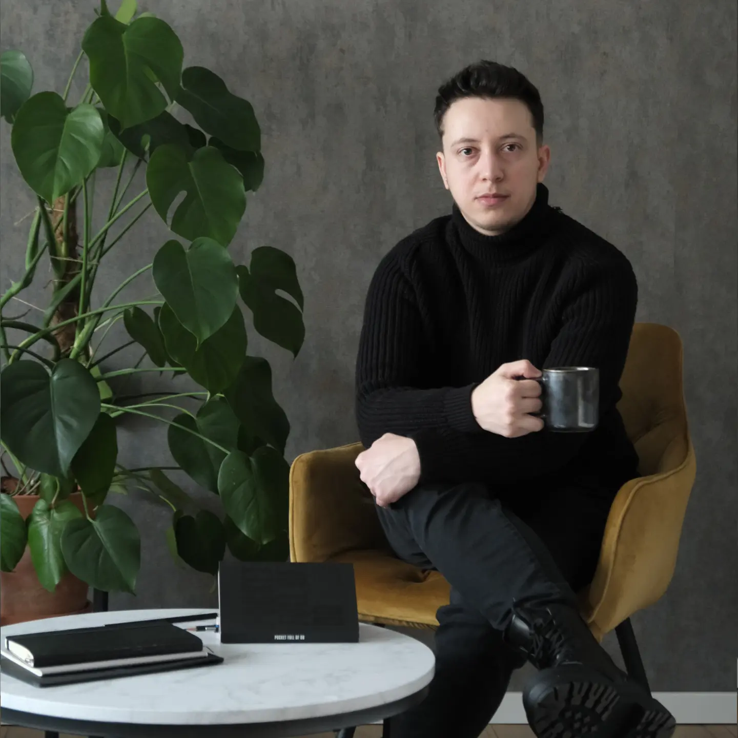 Young man in black sweater sitting on a mustard chair with a thoughtful expression, next to a table with a mug, notebook, and newspaper.